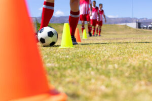 Low angle front view close up of group of Caucasian male football players wearing a team strip, training at a sports field in the sun, waiting their turn to dribble a football between cones, with blue sky in the background